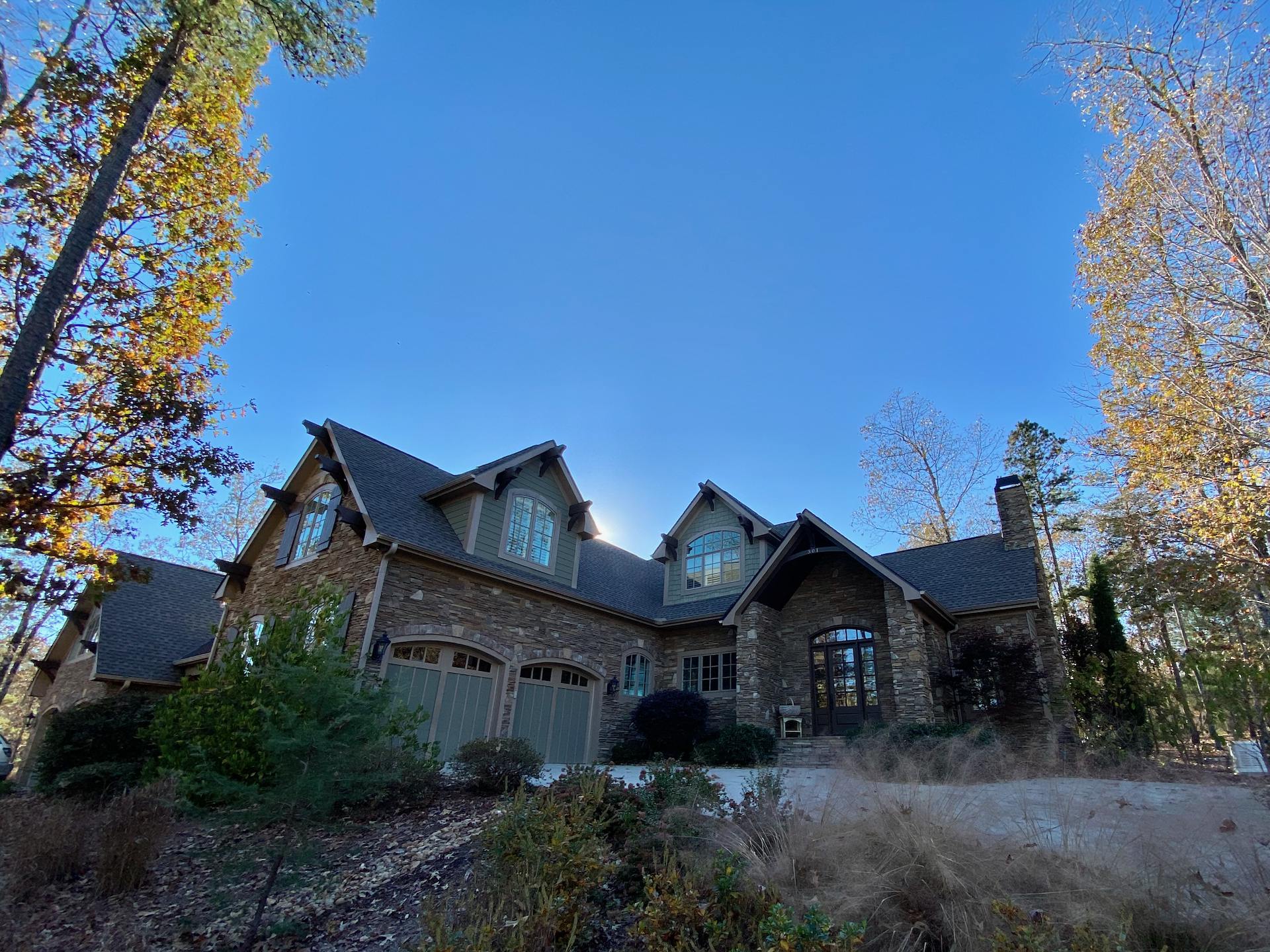 The front and driveway of an L-shaped home surrounded by trees. The facade is natural stone. Two garage doors are on the left side, and two columns surround the arched glass and wood front door on the right.